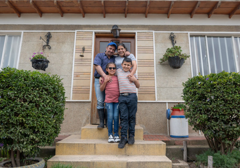 A family of four stands smiling on the steps in front of their home. The parents are standing behind the children, affectionately placing their hands on the children's shoulders. The home features a wooden door, potted plants, and neatly trimmed bushes.