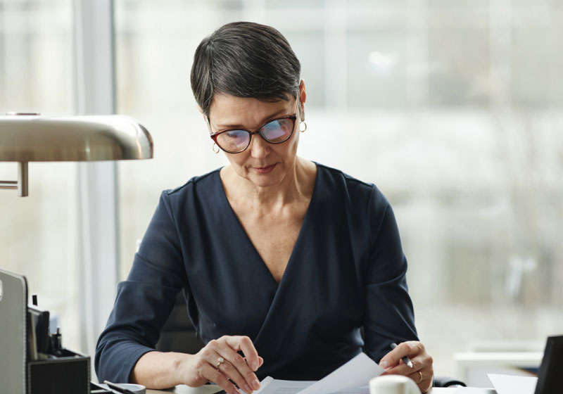 A person with short gray hair and glasses sits at a desk, focused on reading documents. They are wearing a dark blue blouse and are in an office with a window in the background. There's a desk lamp and a cup of coffee beside them.