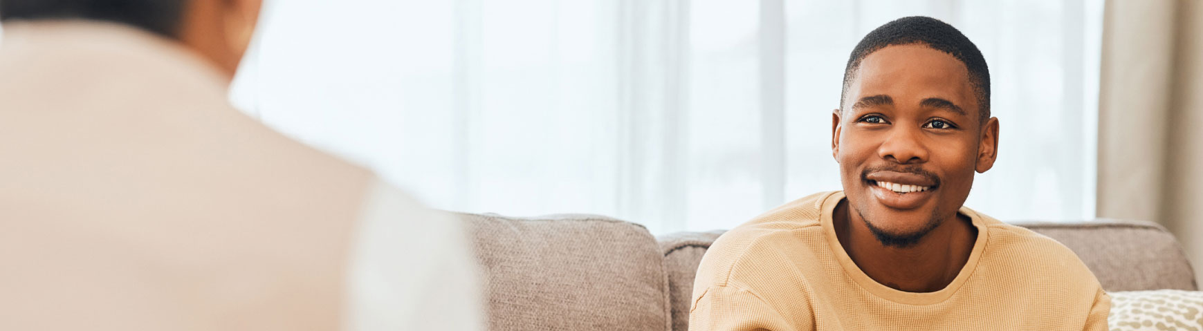 A young man is sitting on a couch, smiling and looking towards another person whose back is to the camera. The background is softly lit, suggesting a comforting, indoor setting.