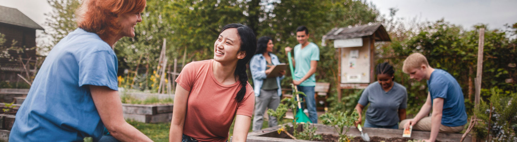 A diverse group of people, including men and women, are working together in a community garden. Some are planting in raised garden beds, while others are chatting and taking notes. The atmosphere is collaborative and the garden is lush with greenery.