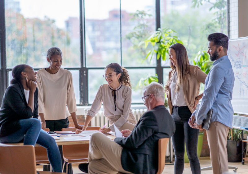 A group of six people are collaborating in an office with large windows and greenery. They are gathered around a table with documents, engaged in discussion, and smiling. One person is seated with papers, while the others stand or lean on the table.