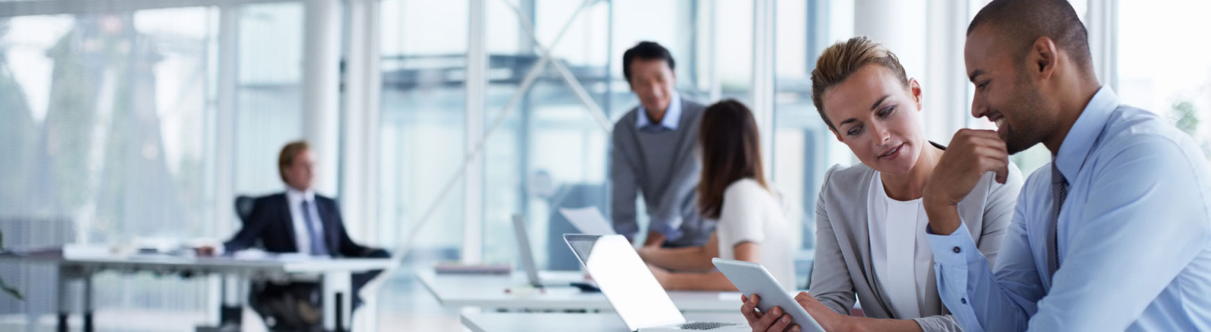 Office setting with five people engaged in various activities. In the foreground, a man and woman discuss something on a tablet. In the background, three people work at desks, with one looking at a laptop and another standing and conversing.