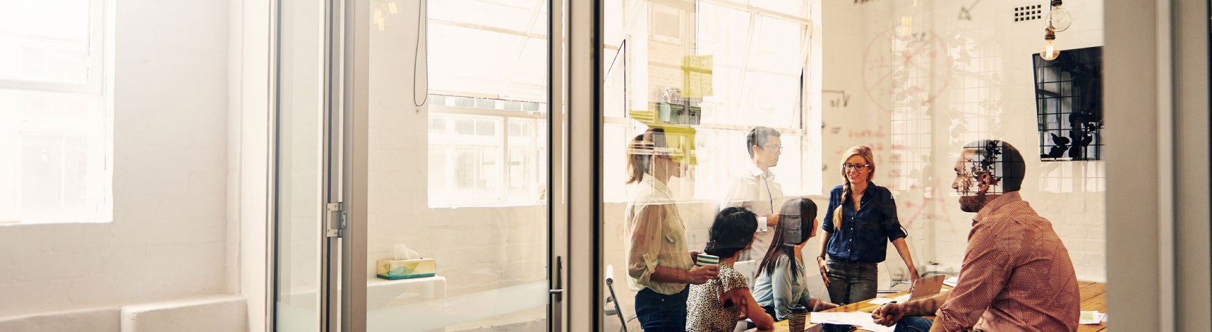 A diverse group of people engaged in conversation and collaboration around a table in a modern office with glass walls and bright windows. Some are seated while others stand, suggesting an informal and dynamic meeting atmosphere.