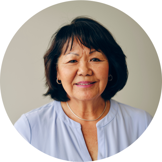 A middle-aged woman with short black hair is smiling at the camera. She is wearing a light purple blouse and a silver necklace. The background is a plain, light-colored wall.