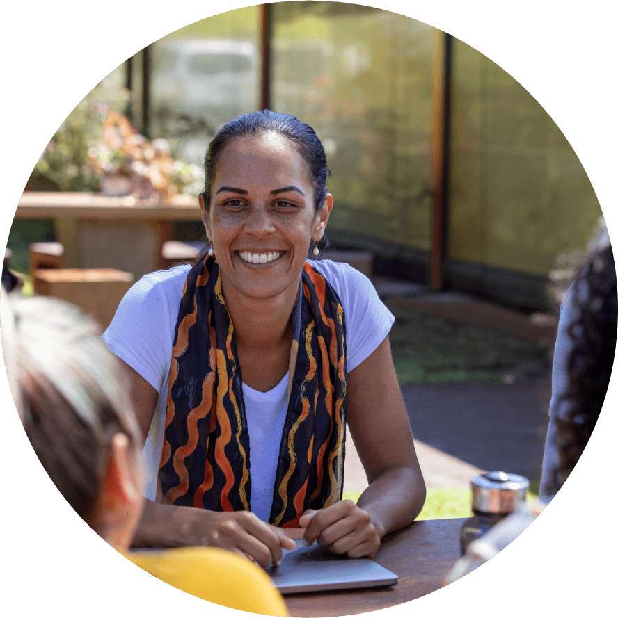 A woman with dark hair tied back, wearing a white shirt and a colorful scarf, is sitting outdoors at a table, smiling and engaging with two other people. The background features greenery and a glass-paneled structure.