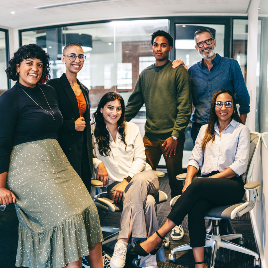 A diverse group of six people smiling and posing in an office setting. Two individuals are seated in office chairs, while the other four stand behind them. The office has large windows, modern furniture, and a collaborative atmosphere.