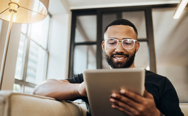 A man with a beard and glasses is sitting on a beige couch in a well-lit room, smiling while looking at a tablet he is holding. There is a lamp and large window in the background, giving the space a cozy and bright feel.