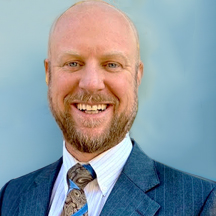 Brian Jones headshot shows him smiling at the camera. He has no hair, a mustache and beard, and wears a blue, pin-stripped suit.