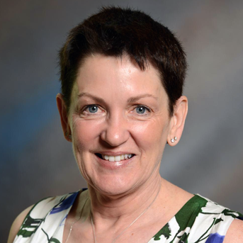 The image shows a headshot of Sherri Downing with short dark hair, smiling. They are wearing a sleeveless top with a green and white floral pattern. The background is a softly blurred gradient of neutral tones.