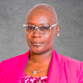 This a headshot of Tamara Mackroy. Tamara has a bald head, wearing glasses, and dressed in a pink jacket over a patterned top. The background is a textured gray.