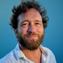 Ari Kirshenbaum, a man with curly hair and a beard is smiling at the camera against a blue background. He is wearing a light-colored, collared shirt.