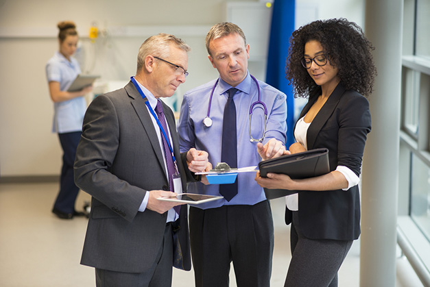 Three medical professionals are in discussion in a bright, modern hospital hallway. One wears a suit, another in a doctor's coat and stethoscope, and a third in business attire holding a tablet. A nurse works in the background.