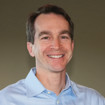 Damien with short, dark hair and a bright smile is wearing a light blue collared shirt. The background is plain and muted.