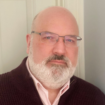 Hugh pictured with glasses and a white beard looking directly at the camera. He is wearing a maroon sweater over a white collared shirt and standing in front of a light-colored wall.