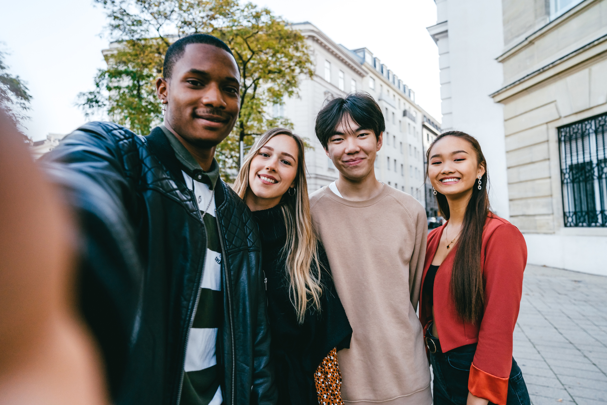 Four friends, including two men and two women, smile for a selfie outdoors in an urban area. They are standing close together, with buildings and trees visible in the background. One person on the left appears to be holding the camera.